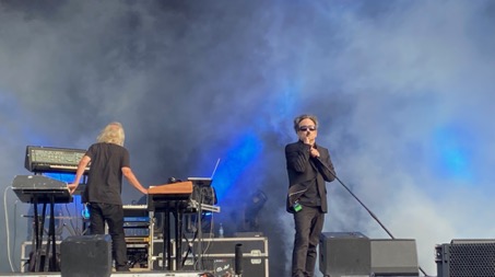 Two musicians dressed in black are standing on a stage. The dark-haired singer wears dark sunglasses and holds a microphone on a microphone stand. The blonde keyboarder stands with his back to the audience and plays two keyboards at the same time. Behind them, a dense, gray fog billows, illuminated in blue. The synthesizers are arranged in a U-shape that is open towards the audience.