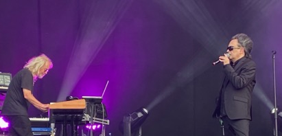 A singer and a keyboard player stand facing each other on a stage. The dark-haired singer is holding a microphone close to his mouth, the blonde keyboarder is looking at the keys he is playing. The stage is bathed in purple light.