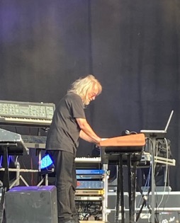 A blonde keyboarder dressed in black is playing on a stage. He is standing to the side of the audience. There are other synthesizers and a laptop around him. The background is black.